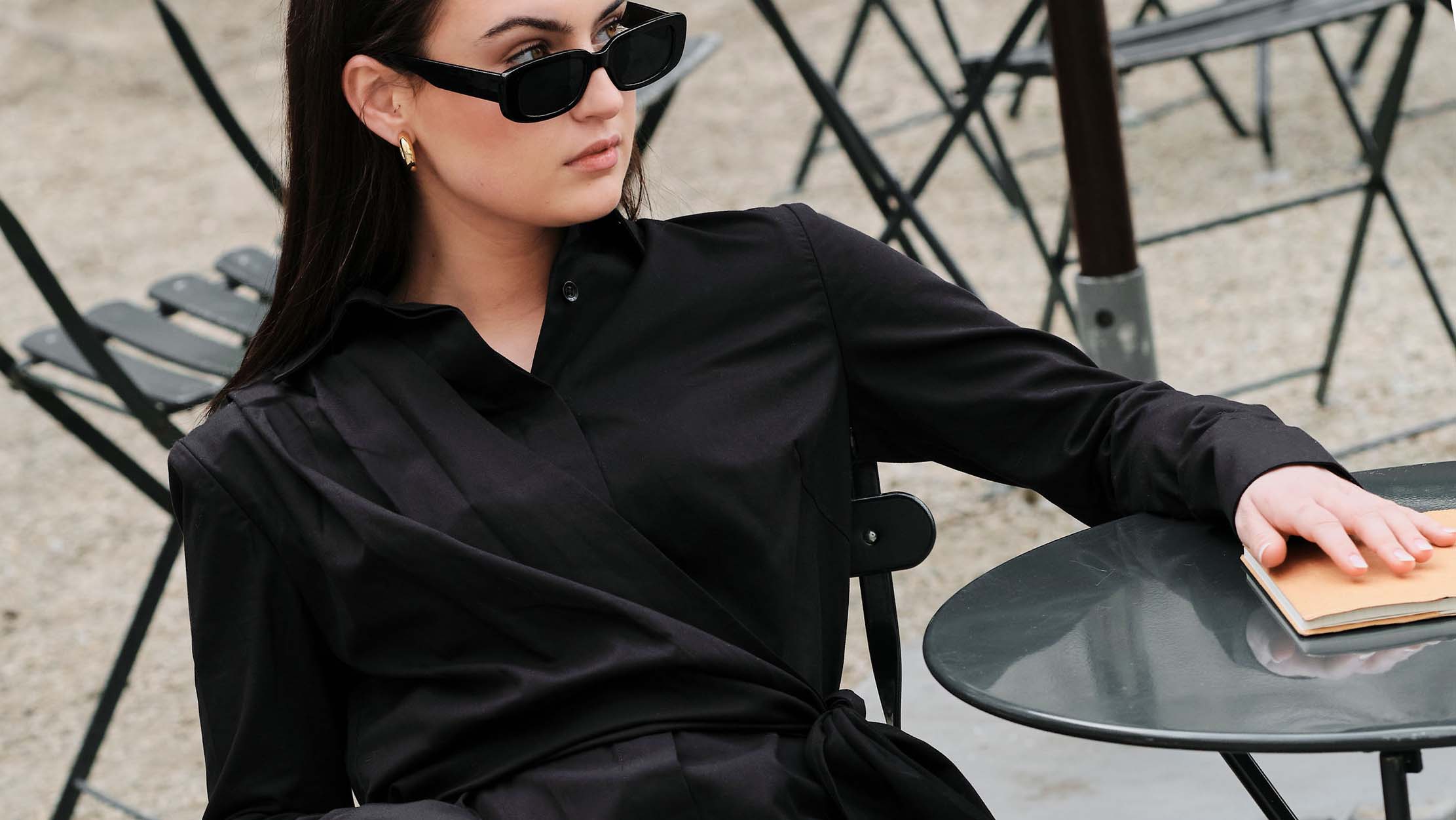 Woman sitting in Bryant Park wearing a black cotton shirt dress by CHABELLA and black sunglasses